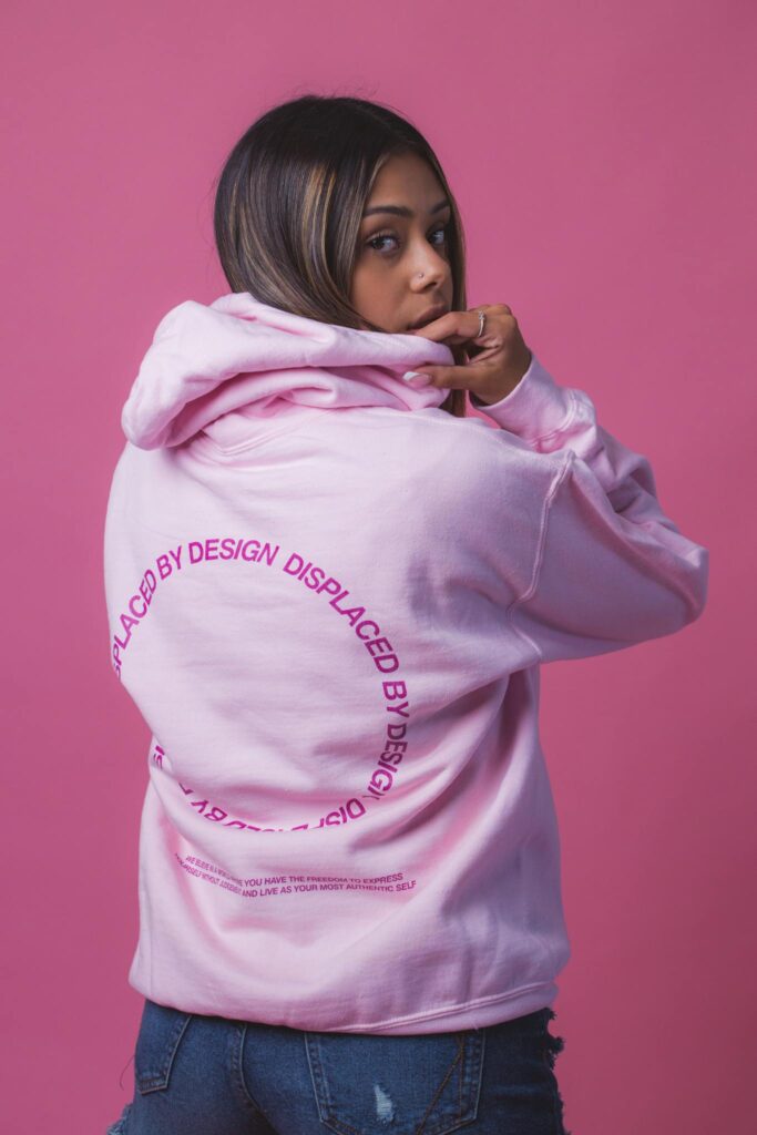 Woman in pink hoodie with 'Displaced by Design' print during studio shoot.