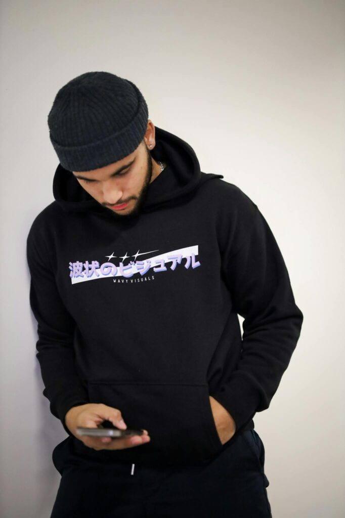 Young man wearing a hoodie and hat, looking down at smartphone indoors.